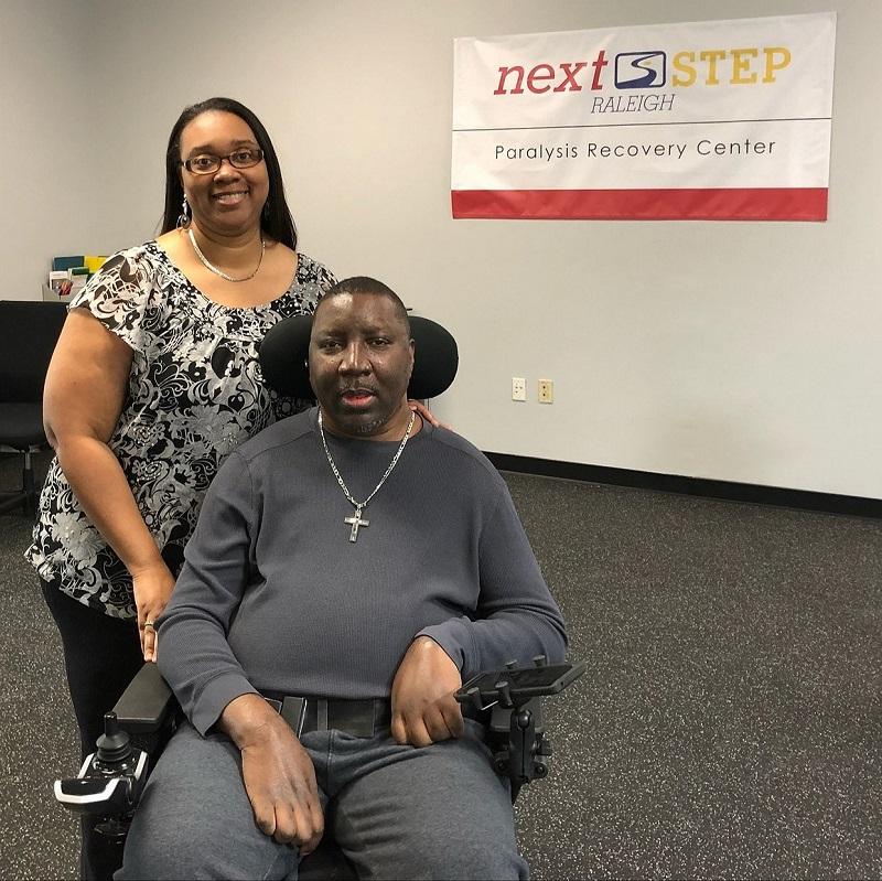 Stephany Golden and his wife Katrina are at Next Step Raleigh for exercise based rehabilitation. Stephany is using a power chair and his wife Katrina is standing beside him.