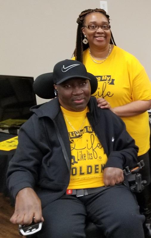 Stephany Golden is wearing a bright yellow fundraising t-shirt and a ball cap and is using a power chair. His wife Katrina is wearing a matching t-shirt and is standing beside him.