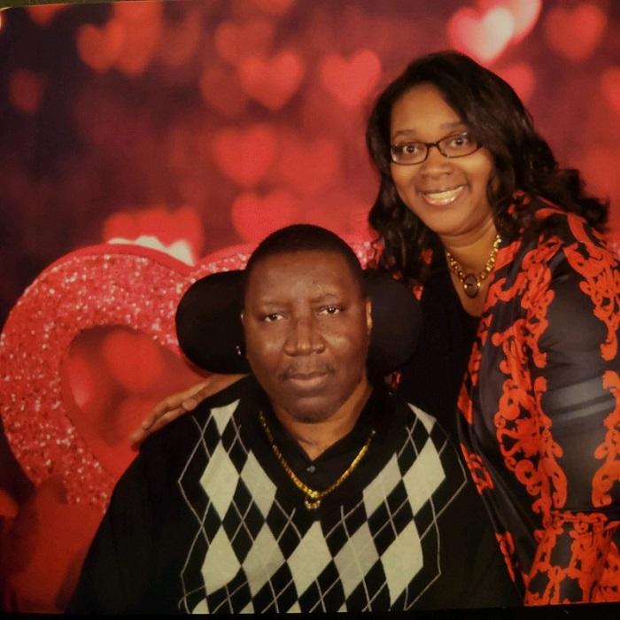 Stephanie and his wife Katrina are pictured with a Valentine's Day backdrop of red and pink hearts. Katrina is standing and smiling at the camera. Stephany is seated in his power chair.