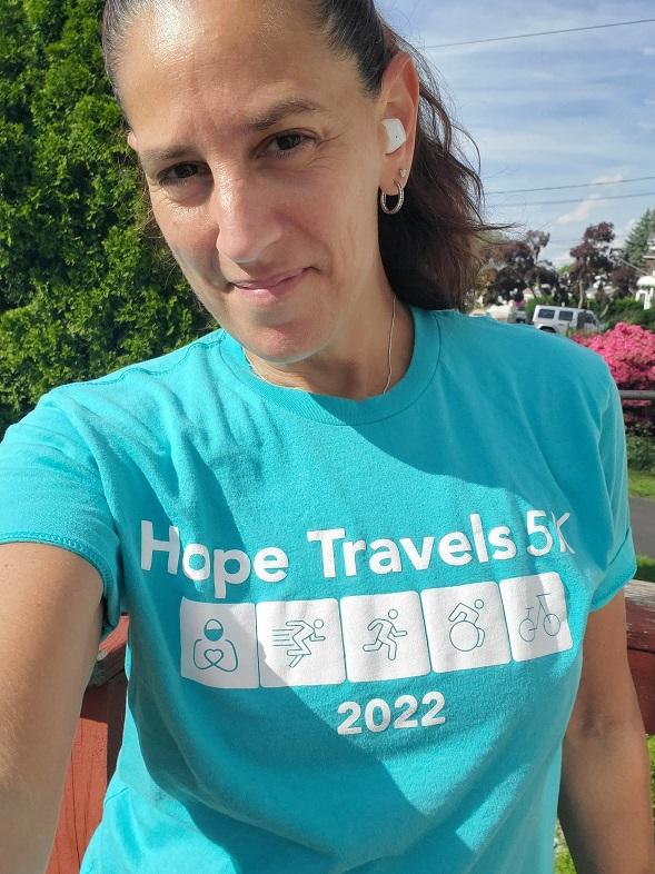A woman smiles at the camera wearing a teal Hope Travels 2022 t-shirt and white airpods. She is outside. She has curly dark brown hair in a ponytail.