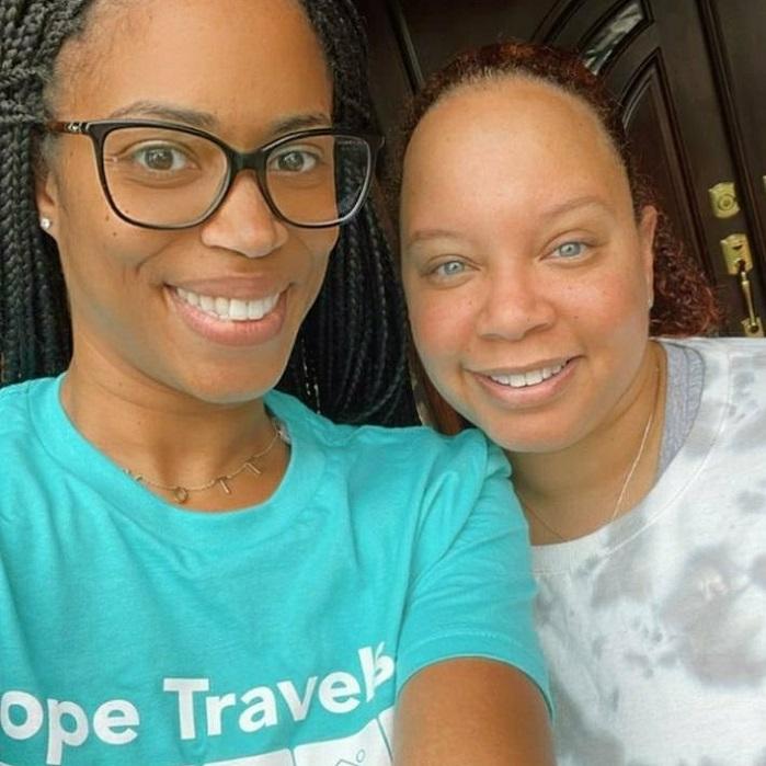 Two women smile at the camera. One has black framed glasses and a teal Hope Travels t-shirt.