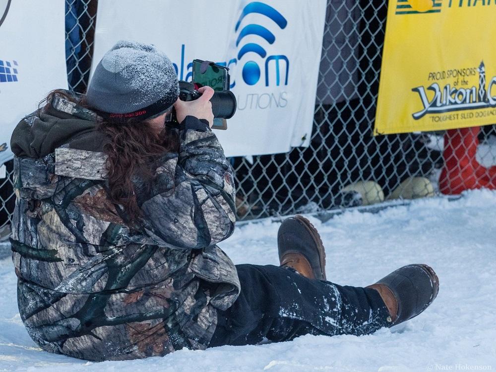 Tracey Porreca is seated in the snow with her legs out in front of her with a large camera in front of her face. She is wearing snow boots, camoflage snow gear, and a gray hat.