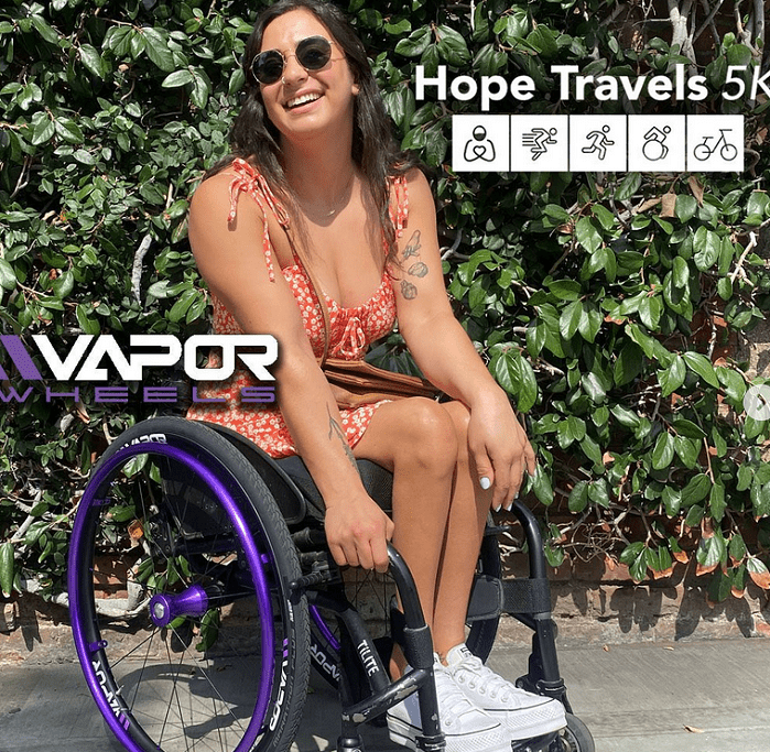 A woman seated in a wheelchair with purple details is wearing sunglasses and an orange and white dress. The Vapor Wheels and Hope Travels 5K logos are visible