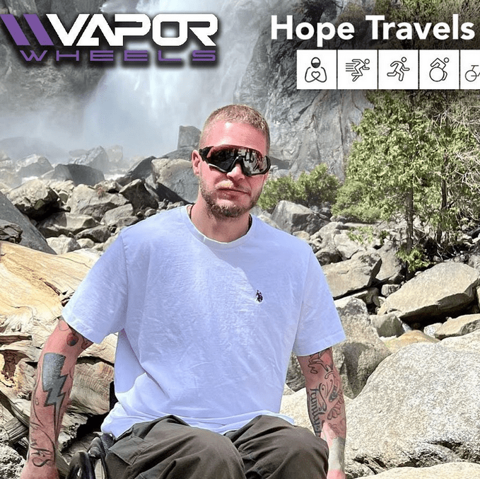 A man seated in his wheelchair is in a white t-shirt, brown pants, and visor-style sunglasses. In the background is a waterfall and a rocky landscape. There are Vapor Wheels and Hope Travels logos.