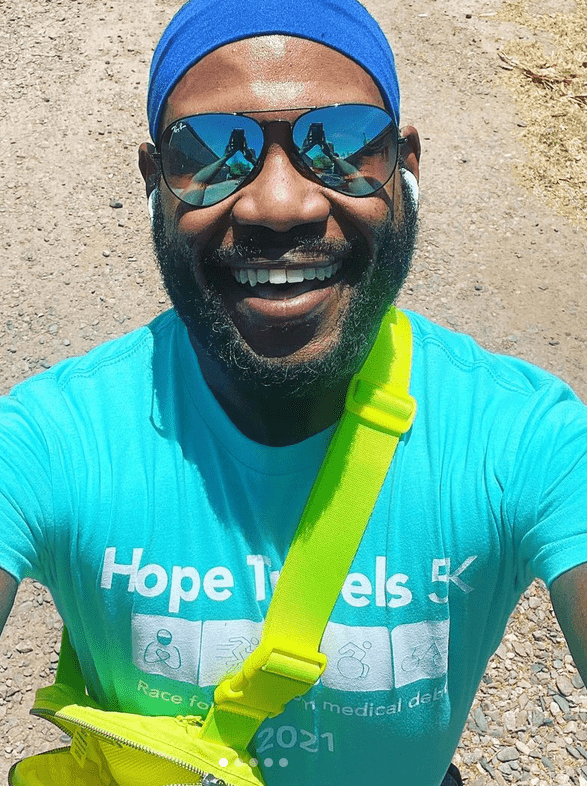 A man smiles at the camera with a teal Hope Travels t-shirt and mirrored sunglasses and Airpods. He has a short curly black beard.