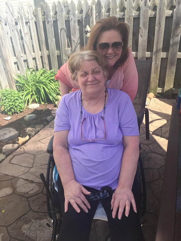 Mary Zagar is seated in her wheelchair with a purple shirt on. She has gray hair. Her daughter Patty is behind her with brown hair, sunglasses, and a pink shirt. They are outside on a patio with a fence.