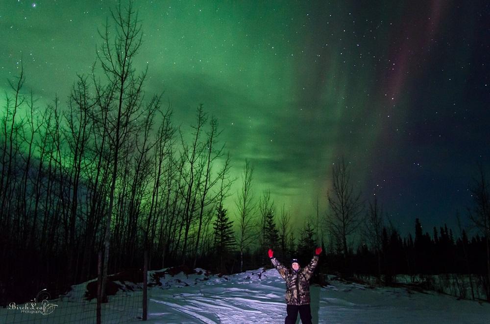 The northern lights are visible in shades of light and dark green behind stark tree silhouettes and white snow. Tracey Porreca is in the foreground with her arms raised and camoflage cold weather gear.