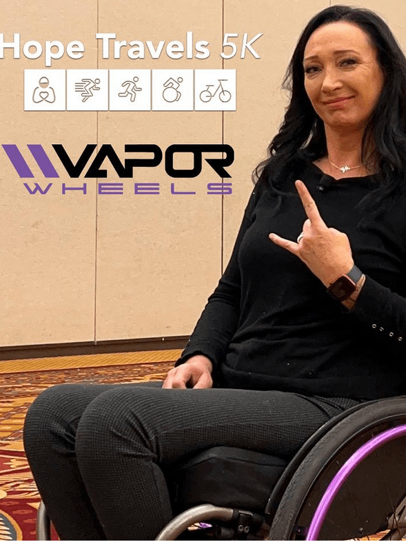 A woman dressed in black is seated in her wheelchair. She has curly dark hair and is giving the rock sign with her right hand. Logos for Vapor Wheels and the Hope Travels Virtual 5K are visible.