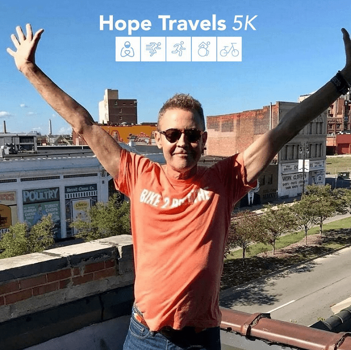 A man stands on a rooftop with a town visible behind him. His arms are outstretched and he wears an orange t-shirt and sunglasses. The Hope Travels 5K logo appears at the top of the picture.