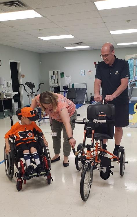 A female presenting person with a nametag points out the adaptive bike’s hockey stick-themed bike to David, who is dressed in Flyers orange and black and is seated next to the bike in his wheelchair. Brad Marsh of the Flyers Alumni Association is holding onto the bike’s back push handles. They are at Schreiber Pediatric Center where David receives therapy.