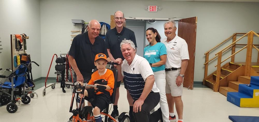 Four members of the Flyers Alumni Association and Help Hope Live Executive Director Kelly L Green stand and kneel near David Albino, a 7-year-old dressed in Flyers themed orange and black, as David sits on his brand-new accessible bike. They are at Schreiber Pediatric Center where David receives therapy.