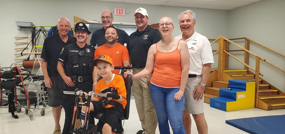 David is seated on his new adaptive bike dressed in Flyers orange and black with a big smile. Behind him are members of the Flyers Alumni Association, his mother in an orange tank top with a bald head and black-framed glasses, and a uniformed officer. Everyone is laughing and smiling. They are at Schreiber Pediatric Center where David receives therapy.