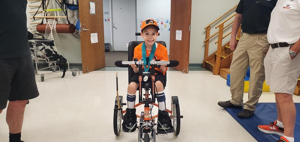 7-year-old David Albino is seated on his new adaptive tricycle wearing Philadelphia Flyers orange and black clothing. He is smiling. He is inside of Schreiber Pediatric Center where he receives therapy.