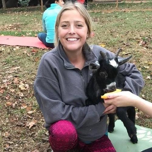 Female presenting multi-visceral transplant recipient Catherine is smiling wide wearing a gray athletic sweater and bright pink athletic pants. She has blonde hair framing her gace and dark eyes with light tan skin. She is holding a black baby goat.