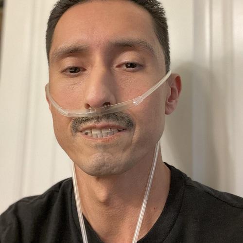 Male presenting double lung transplant recipient Jair has close cropped dark hair and dark eyes with a light mustache. He is wearing an oxygen tube.
