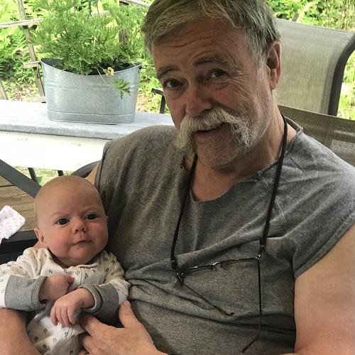 Male presenting double lung transplant recipient Jay has light colored skin and is wearing a gray shirt with short sleeves and a pair of glasses around his neck. He has gray hair, dark eyes, and a gray handlebar mustache. He is holding a small baby in his right arm.