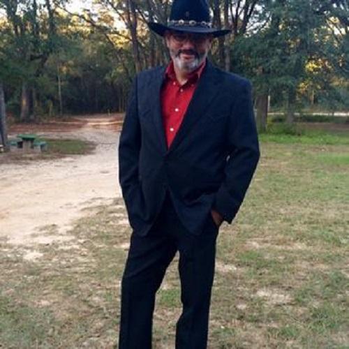 Male presenting heart transplant recipient John is outdoors wearing a full dark blue suit, a red shirt, and a matching blue cowboy hat. His silver facial hair is visible.