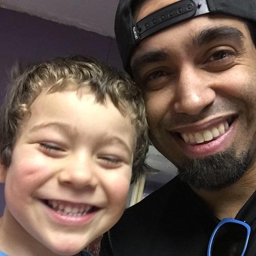 Male presenting kidney transplant recipient Lafe Timothy has medium brown skin, black facial hair, and dark eyes and is wearing a backward black ball cap. To his right is his young son who has lighter colored skin and hair and a big smile.