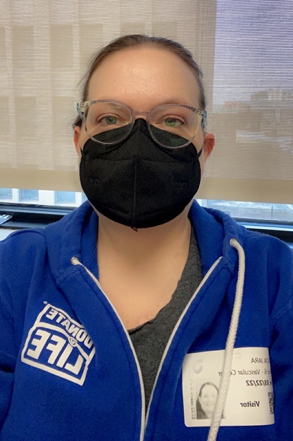 Linda Jara is wearing gray frame glasses and a black mask wit her hair pulled back. She has green eyes and is wearing a blue Donate Life hoodie with white details in an indoor setting.