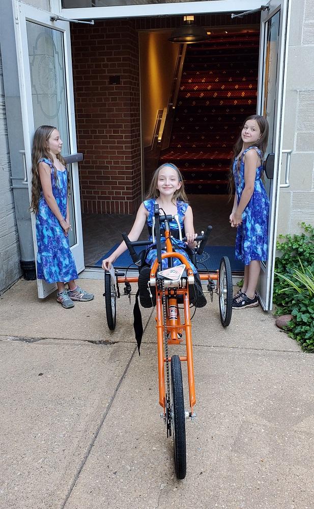 10-year-old Madalynn Sheerin is seated on her new orange Freedom Concepts handcycle wearing a blue and light blue tie-dye dress. Behind her, her two sisters are wearing matching dresses and sneakers and are holding open the doors to a venue. All three have long brown hair.