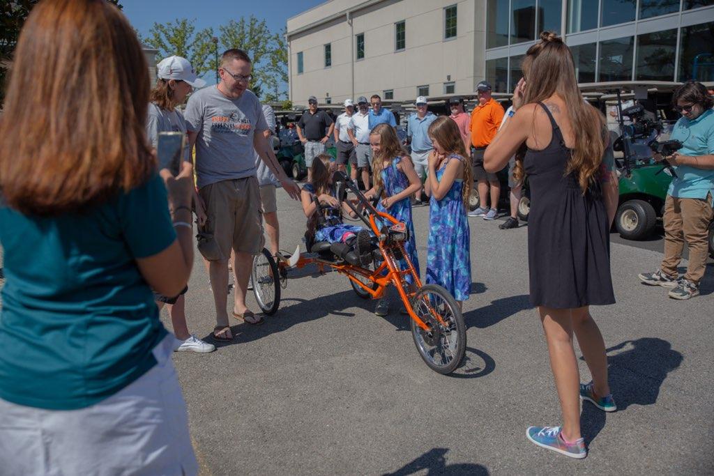 Madalynn Sheerin is seated on her new orange handcycle in an outdoor parking lot. Next to her are her two sisters. All three have long brown hair and are wearing blue and light blue tie-dye dresses. Family members, friends, and other Flyers Golf Outing attendees are standing nearby with golf carts visible in the background.