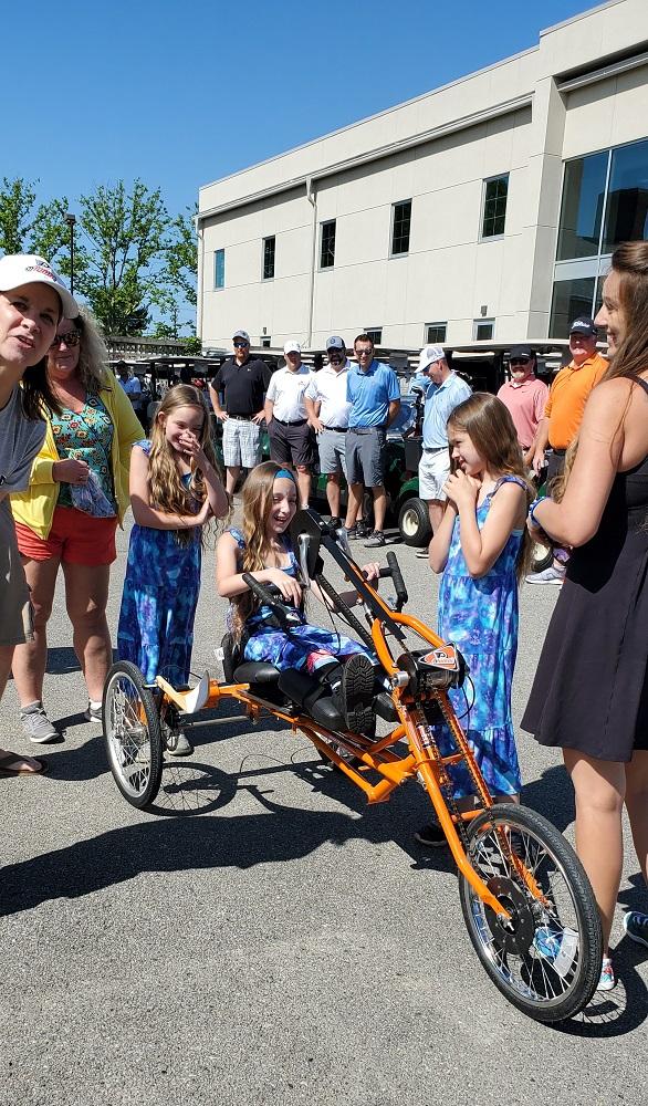 Madalynn rides her new orange Flyers-themed handcycle with her two sisters walking beside her. All three have long brown hair and are wearing blue and light blue tie-dye dresses. It’s a clear and sunny day. Behind them, a row of individuals in golf attire applaud in front of a row of golf carts.