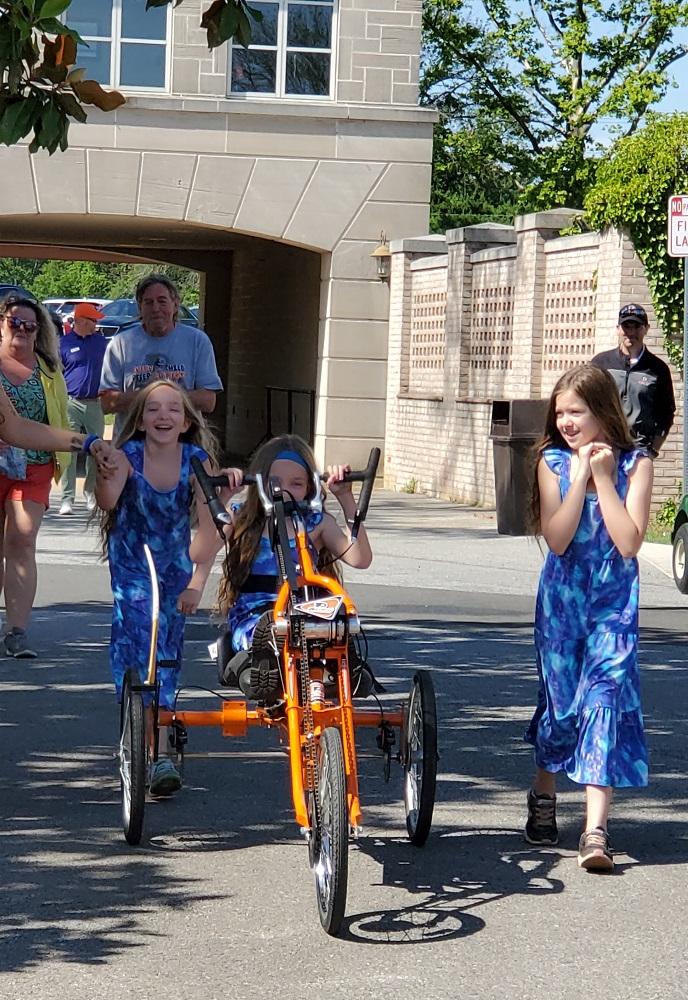 Madalynn excitedly uses her new orange handcycle with her two sisters walking along with her and smiling. All three have long brown hair and are wearing blue and light blue tie-dye dresses.