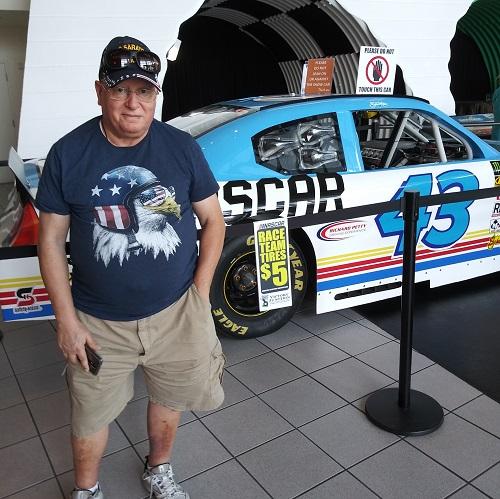 Liver transplant recipient Salvatore is a middle aged male presenting individual wearing a navy blue t-shirt with an American bald eagle on it and a pair of glasses. Behind him is a NASCAR car on display.