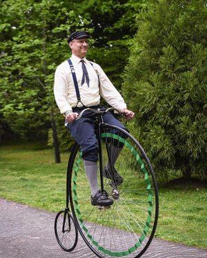 Bill Soloway sits atop his high wheel historic bicycle wearing old-fashioned high gray socks, suspenders, a long flowing bow tie, and a peddler's cap.