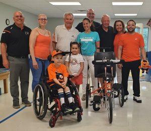 7-year-old David is in his wheelchair in a Philadelphia Flyers orange and black jersey and ball cap. His family, the Flyers Alumni Association, and Help Hope Live’s Executive Director are standing behind him and smiling. To his left is his new Flyers themed orange and black adaptive bike from Freedom Concepts. They are at Schreiber Pediatric Center where David receives therapy. Blue exercise mats are visible.