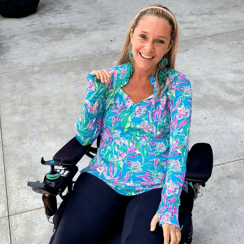 Ali Ingersoll is radiant in a brightly-colored floral print long-sleeved shirt with a tan headband over her blonde hair and a big smile. Living with paralysis, Ali is seated in a black power chair.