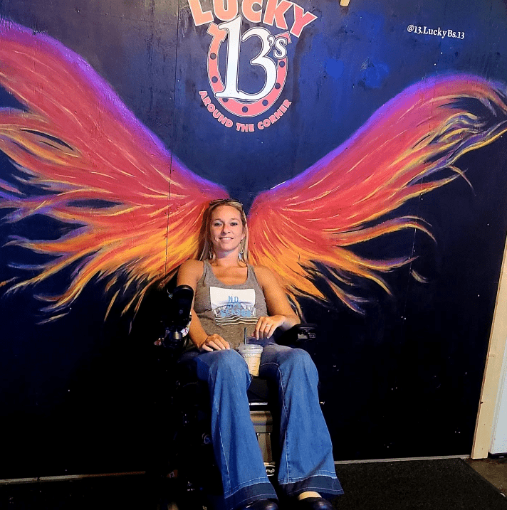 Ali Ingersoll, a woman living with paralysis, is in her black power chair in flared jeans and an olive tank top. She is in front of a wall that features a midnight blue background with brightly-colored painted wings that appear to be coming out of her back. A logo above the large wings reads Lucky 13s, likely the name of the establishment.