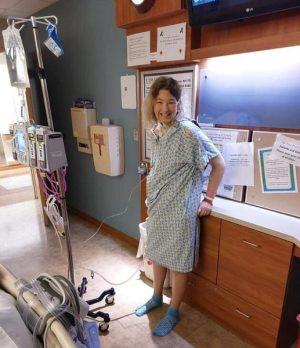 Kidney transplant recipient Jennifer Kershner is pictured in a hospital room wearing a hospital gown and hospital socks. She is smiling with white skin and shoulder length brown and blonde hair. She is hooked up to a rolling IV cart.