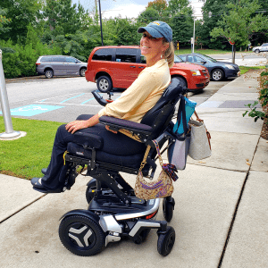 Ali Ingersoll, a woman living with paralysis, is seated in a black adjustable electric wheelchair. She is wearing a yellow shirt, earrings, and a blue ball cap with a ring on her left ring finger.
