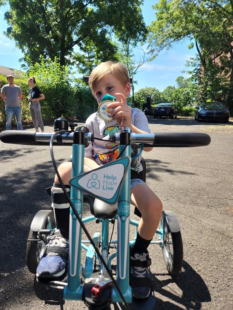 Vinny holds up his Help Hope Live themed medal for the camera, covering his nose and mouth. He is four years old with light skin and blonde hair with brown eyes. He is wearing a gray t-shirt and jean shorts as he sits on his teal and white Help Hope Live themed adaptive bike. He is wearing a black leg brace on his lower right leg. Behind him is a blacktop with trees and a blue sky visible.