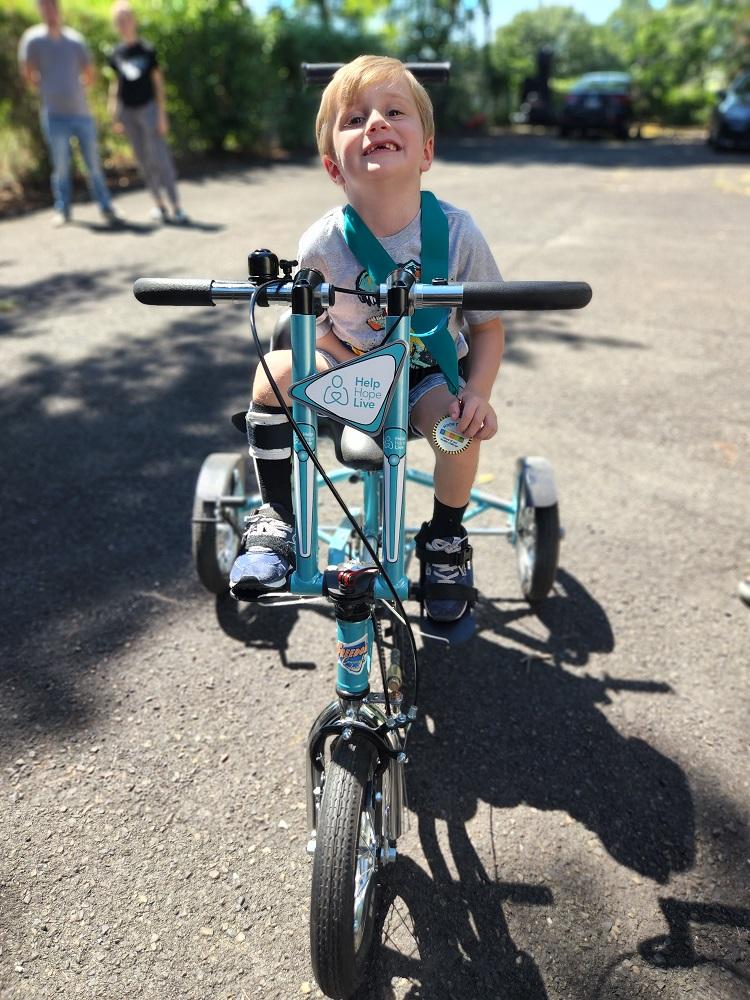 4-year-old Vinny sits on his teal and white Help Hope Live themed adaptive bike and smiles wide. He is wearing a black leg brace on his lower right leg. Behind him is a blacktop with trees and a blue sky visible.