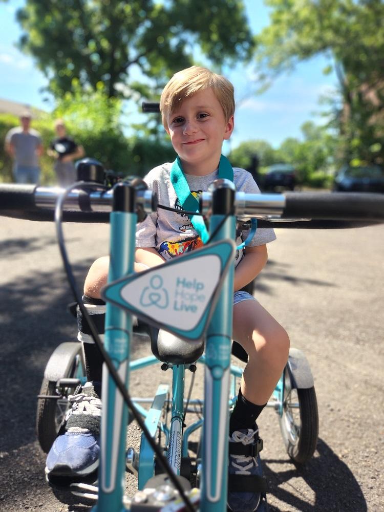 4-year-old Vinny sits on his teal and white Help Hope Live themed adaptive bike. He is wearing a black leg brace on his lower right leg. Behind him is a blacktop with trees and a blue sky visible.