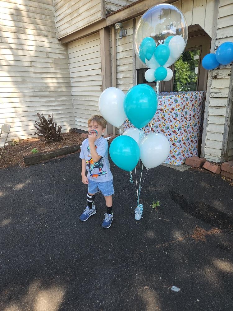 On the blacktop in front of his home, 4-year-old Vinny wears a t-shirt and shorts along with a black brace on his right lower leg. He hides his smile behind a teal Help Hope Live wristband. Next to him are teal and white balloons, and behind him is a large wrapped box.
