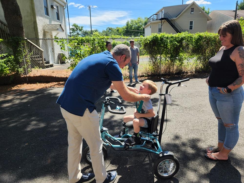 4-year-old Vinny receives a Help Hope Live themed medal on a teal fabric neck strap as he sits on his Freedom Concepts adaptive bike. He is outside with his family.