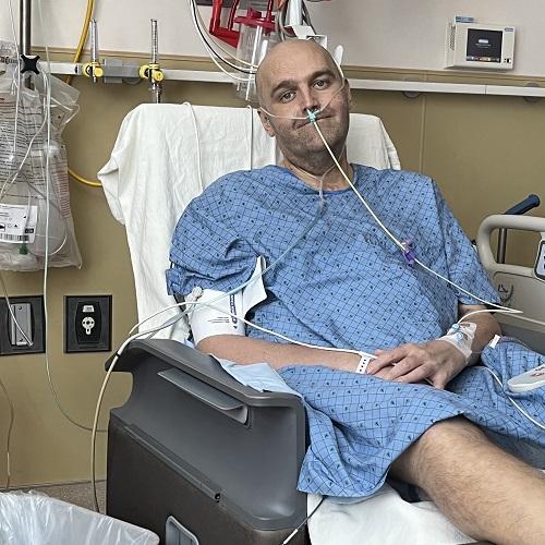Liver transplant recipient Brent is in the hospital seated wearing a blue hospital gown with a shaved head and light skin. He is hooked up to several machines.