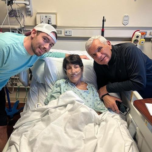 Double lung transplant recipient Cathy is pictured in the hospital wearing a hospital gown. She has short dark hair and light skin. Beside her are two male presenting supporters, one with light brown facial hair and a white backwards baseball cap and one with white hair and a blue turtleneck.