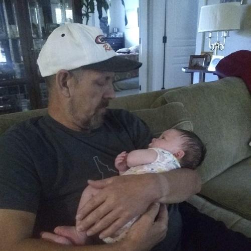 Liver transplant recipient Dale holds a baby. He has light skin and a white baseball cap.