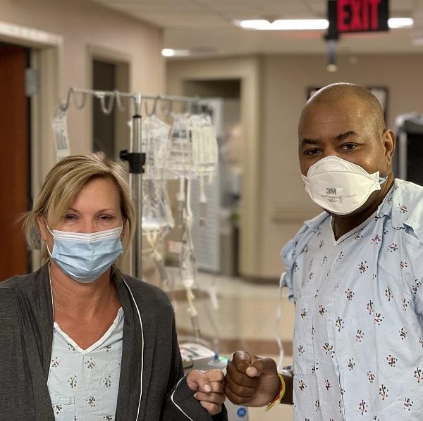 Keith Henry wears a hospital gown and a white mask and bumps fists with his living donor, also in a hospital gown. Keith has brown skin and a bald head and his living donor has blonde hair and light skin (female-presenting).