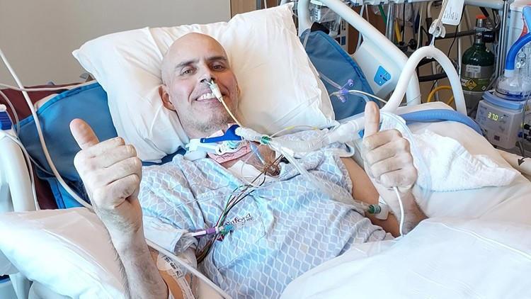 Steven Whitaker is pictured in a hospital setting with a hospital gown and many tubes connected to his body and nose. He has light skin, a bald head, and a smile and he's giving two thumbs up.