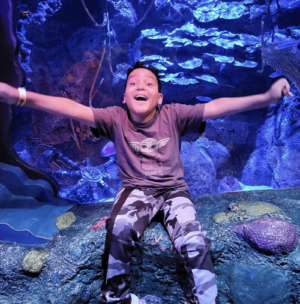 Kelson wears a brown shirt with a baby yoda on it and gray camoflage print pants as he poses in front of an aquarium. He has a huge smile and is extending his arms to the left and right in excitement. He has light brown skin, curly dark hair, and dark eyes.