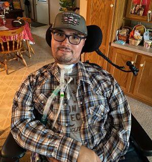 Derek Szena wears black rimmed glasses, a gray ball cap, and a plaid shirt and uses a ventilator as he sits in his black power chair. He has light skin with a faint black goatee.
