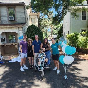 4-year-old Vinny wears a leg brace on his lower right leg as he sits on his new teal adaptive bike from Help Hope Live, which is themed to match the nonprofit’s color scheme and logo. Behind him is his family, Help Hope Live supporters, and Help Hope Live Executive Director Kelly L Green wearing a Help Hope Live teal t-shirt. There are teal and white balloons beside them. They are on a neighborhood blacktop directly in front of Vinny’s home with a blue sky visible.