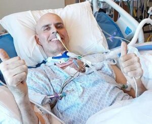 Steven Whitaker is pictured in a hospital setting with a hospital gown and many tubes connected to his body and nose. He has light skin, a bald head, and a smile and he's giving two thumbs up.