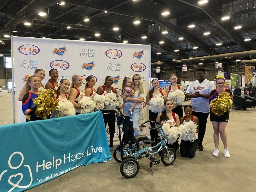 At the Abilities Expo Phoenix, 3-year-old Emberly and her mom are surrounded by cheerleaders as they stand behind Emberly’s new adaptive bike.