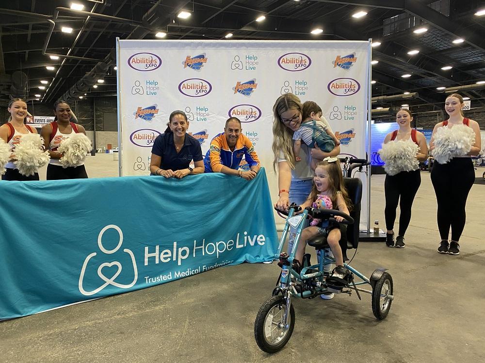 James from Freedom Concepts and Kelly L Green from Help Hope Live look on and smile as Emberly grins on her new adaptive bike. Her mom holds one side of the handlebars above her. They are at the Abilities Expo Phoenix next to a large Help Hope Live teal banner with cheerleaders nearby.